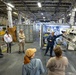 A group of civilians and military/uniformed personnel are gathered inside what appears to be the hangar bay or interior deck of a large naval vessel or military facility. A uniformed individual is conducting a briefing or tour, pointing toward large white cylindrical equipment mounted on a mechanical rack or launcher system. The setting features industrial infrastructure including insulated pipes, overhead lighting, and shipping containers.