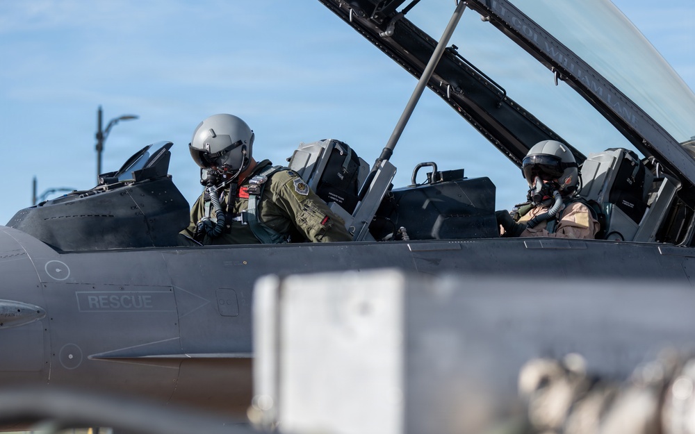 Two military pilots are seated in the tandem cockpit of a fighter jet, both wearing full flight gear including helmets and oxygen masks. The aircraft fuselage displays the word 'RESCUE' near the cockpit area, consistent with standard military aircraft safety markings. The image appears to be taken on a flight line or tarmac under clear blue sky conditions.