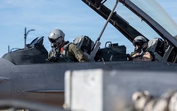 Two military pilots are seated in the tandem cockpit of a fighter jet, both wearing full flight gear including helmets and oxygen masks. The aircraft fuselage displays the word 'RESCUE' near the cockpit area, consistent with standard military aircraft safety markings. The image appears to be taken on a flight line or tarmac under clear blue sky conditions.