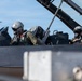 Two military pilots are seated in the tandem cockpit of a fighter jet, both wearing full flight gear including helmets and oxygen masks. The aircraft fuselage displays the word 'RESCUE' near the cockpit area, consistent with standard military aircraft safety markings. The image appears to be taken on a flight line or tarmac under clear blue sky conditions.