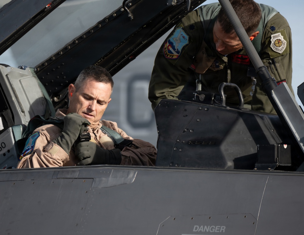 Two military personnel are shown at a fighter jet cockpit, with one pilot seated inside the aircraft and another individual in a flight suit leaning over to assist or brief him. The seated pilot is wearing a tan/desert flight suit with patches and appears to be adjusting his harness or equipment. The word 'DANGER' is visible on the aircraft fuselage, consistent with standard military aircraft safety markings.