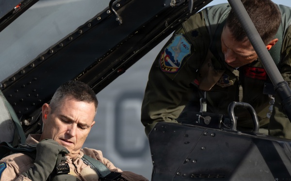 Two military personnel are shown at a fighter jet cockpit, with one pilot seated inside the aircraft and another individual in a flight suit leaning over to assist or brief him. The seated pilot is wearing a tan/desert flight suit with patches and appears to be adjusting his harness or equipment. The word 'DANGER' is visible on the aircraft fuselage, consistent with standard military aircraft safety markings.