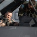 Two military personnel are shown at a fighter jet cockpit, with one pilot seated inside the aircraft and another individual in a flight suit leaning over to assist or brief him. The seated pilot is wearing a tan/desert flight suit with patches and appears to be adjusting his harness or equipment. The word 'DANGER' is visible on the aircraft fuselage, consistent with standard military aircraft safety markings.
