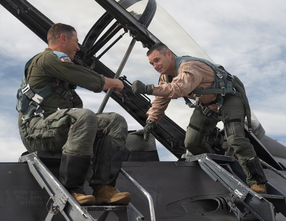 Two military aviators are interacting on top of a fighter jet, appearing to shake hands or exchange something near the cockpit area. Both are wearing flight gear including harnesses, G-suits, and flight suits of different colors — one in olive green and one in tan/khaki. The setting appears to be an outdoor airfield with a partly cloudy sky in the background.