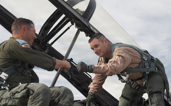 Two military aviators are interacting on top of a fighter jet, appearing to shake hands or exchange something near the cockpit area. Both are wearing flight gear including harnesses, G-suits, and flight suits of different colors — one in olive green and one in tan/khaki. The setting appears to be an outdoor airfield with a partly cloudy sky in the background.