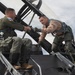 Two military aviators are interacting on top of a fighter jet, appearing to shake hands or exchange something near the cockpit area. Both are wearing flight gear including harnesses, G-suits, and flight suits of different colors — one in olive green and one in tan/khaki. The setting appears to be an outdoor airfield with a partly cloudy sky in the background.