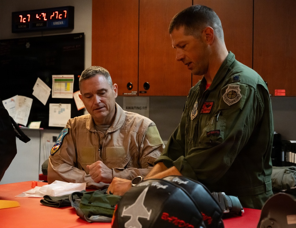 Two military aviators, one in a tan/khaki flight suit and one in an olive green flight suit, are reviewing documents or paperwork at a red-topped table in what appears to be a flight operations or equipment room. Both wear military patches on their uniforms, and flight equipment including a helmet bag is visible on the table. A digital clock display showing LOCAL, ZULU, and ARENA times is visible in the background.