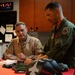 Two military aviators, one in a tan/khaki flight suit and one in an olive green flight suit, are reviewing documents or paperwork at a red-topped table in what appears to be a flight operations or equipment room. Both wear military patches on their uniforms, and flight equipment including a helmet bag is visible on the table. A digital clock display showing LOCAL, ZULU, and ARENA times is visible in the background.