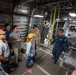 A group of civilians is receiving a guided tour inside what appears to be a military vessel or aircraft, likely a US Coast Guard or Navy ship. Two personnel in uniform coveralls are conducting the briefing, explaining equipment to approximately 8-10 visitors. The interior shows industrial metallic construction with mechanical equipment including a large winch or cable reel system.