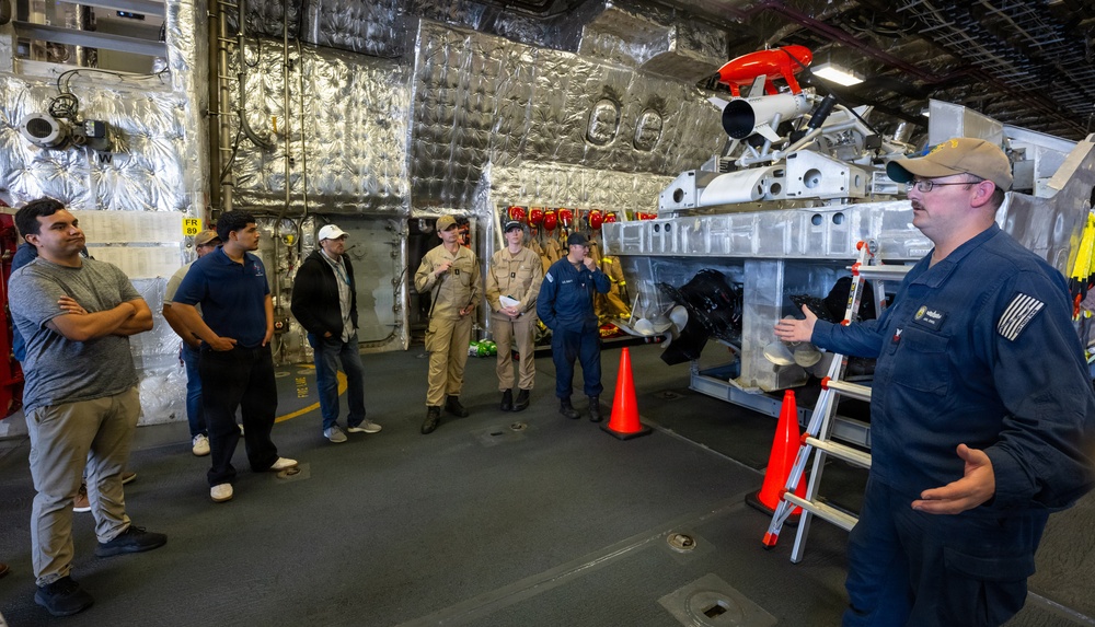 A uniformed personnel member, likely U.S. Coast Guard based on visible insignia and uniform style, is briefing a group of civilians and uniformed colleagues inside what appears to be a ship's hangar bay or equipment bay. The space contains a large piece of machinery or equipment, possibly a remotely operated vehicle (ROV) or specialized maritime equipment, with a red component visible on top. Safety cones and a ladder are positioned near the equipment.