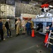 A uniformed personnel member, likely U.S. Coast Guard based on visible insignia and uniform style, is briefing a group of civilians and uniformed colleagues inside what appears to be a ship's hangar bay or equipment bay. The space contains a large piece of machinery or equipment, possibly a remotely operated vehicle (ROV) or specialized maritime equipment, with a red component visible on top. Safety cones and a ladder are positioned near the equipment.