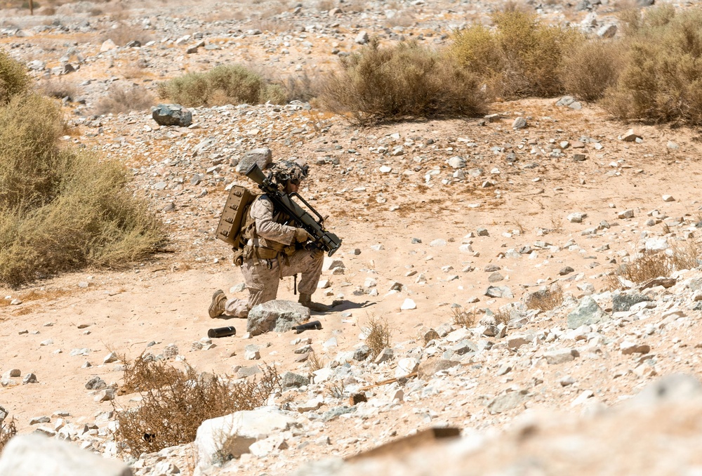 A single soldier in desert camouflage uniform is kneeling on rocky, arid terrain in a tactical position. The individual is heavily equipped with a backpack, helmet, and what appears to be a shoulder-fired weapon system. The environment is consistent with a desert training or operational area characterized by sparse scrub brush and rocky ground.
