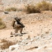 A single soldier in desert camouflage uniform is kneeling on rocky, arid terrain in a tactical position. The individual is heavily equipped with a backpack, helmet, and what appears to be a shoulder-fired weapon system. The environment is consistent with a desert training or operational area characterized by sparse scrub brush and rocky ground.