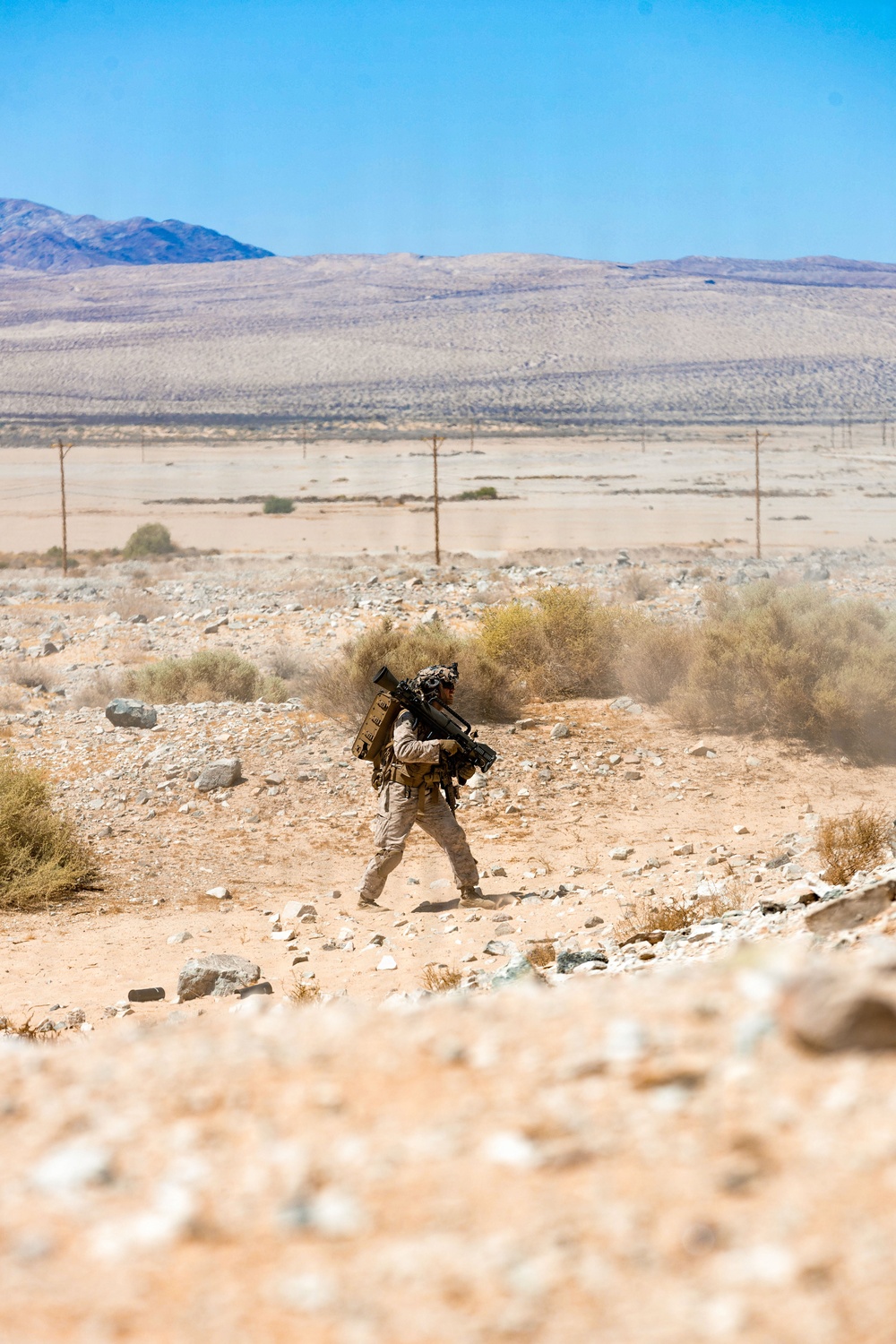 A single soldier in desert camouflage uniform moves across arid terrain carrying what appears to be a shoulder-fired weapon system (likely an AT4 or similar rocket launcher) on their back along with additional equipment. The setting is a stark desert landscape with sparse scrub vegetation, rocky ground, utility poles in the mid-distance, and mountain ridgelines visible on the horizon under a clear blue sky. The image appears to be taken during a military training exercise in a desert environment consistent with locations such as the Mojave Desert.