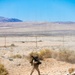 A single soldier in desert camouflage uniform moves across arid terrain carrying what appears to be a shoulder-fired weapon system (likely an AT4 or similar rocket launcher) on their back along with additional equipment. The setting is a stark desert landscape with sparse scrub vegetation, rocky ground, utility poles in the mid-distance, and mountain ridgelines visible on the horizon under a clear blue sky. The image appears to be taken during a military training exercise in a desert environment consistent with locations such as the Mojave Desert.