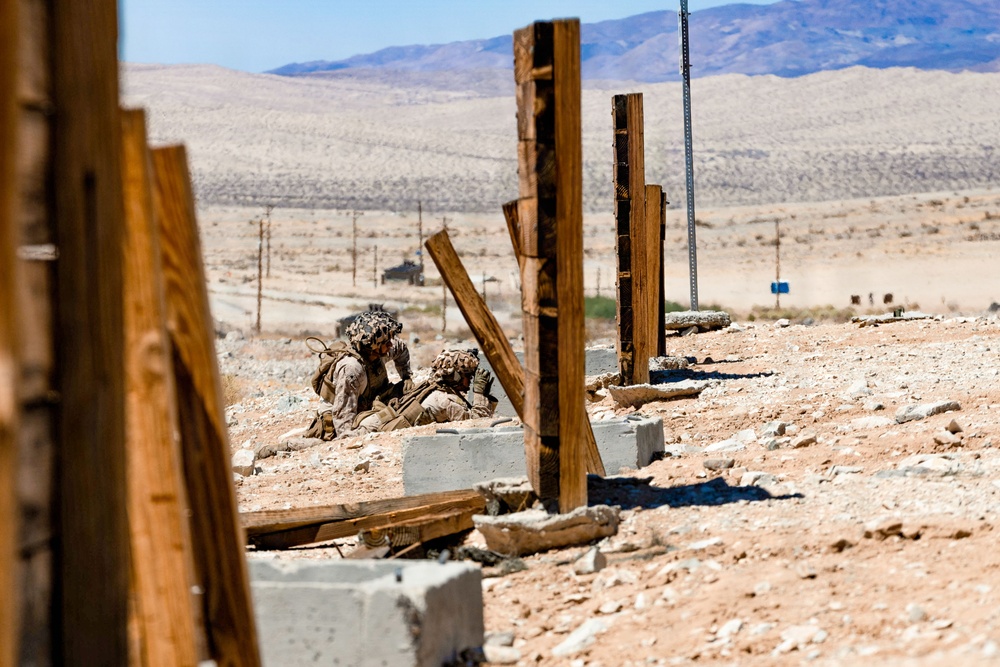 Two military personnel in desert camouflage uniforms are taking cover behind concrete blocks and wooden structures in an arid desert training environment. The setting appears to be a military training facility with simulated urban rubble, wooden post frameworks, and open desert terrain stretching to distant mountains in the background. A military vehicle is faintly visible in the mid-distance along a road.