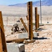 Two military personnel in desert camouflage uniforms are taking cover behind concrete blocks and wooden structures in an arid desert training environment. The setting appears to be a military training facility with simulated urban rubble, wooden post frameworks, and open desert terrain stretching to distant mountains in the background. A military vehicle is faintly visible in the mid-distance along a road.