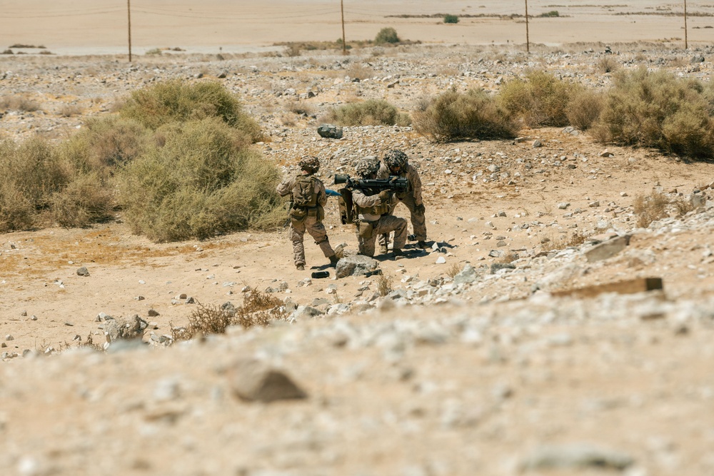 Two soldiers in desert camouflage uniforms are operating in an arid, rocky terrain with sparse desert scrub vegetation. One soldier appears to be kneeling and aiming a large shoulder-mounted weapon system while the other provides assistance or support. The setting suggests a desert environment, consistent with a training exercise or operational deployment.