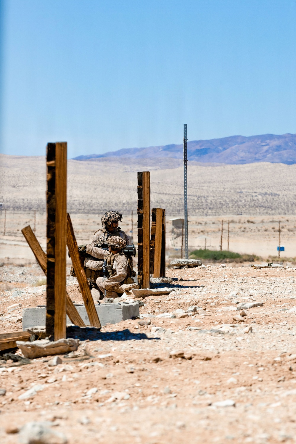 Two military personnel in desert camouflage uniforms are taking cover behind wooden post structures in an arid desert training environment. They appear to be conducting a tactical exercise, with one soldier aiming a weapon while the other observes. The setting features rocky, sandy terrain with distant mountains and a clear blue sky.