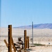 Two military personnel in desert camouflage uniforms are taking cover behind wooden post structures in an arid desert training environment. They appear to be conducting a tactical exercise, with one soldier aiming a weapon while the other observes. The setting features rocky, sandy terrain with distant mountains and a clear blue sky.