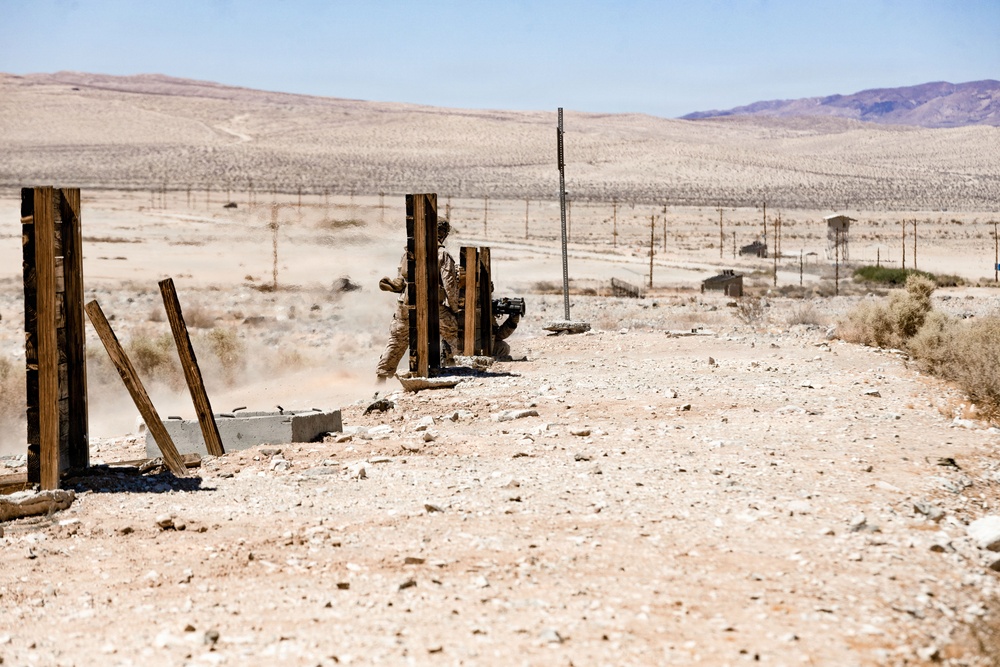 A soldier in desert camouflage uniform is positioned between wooden posts in an arid desert training environment, appearing to be in a tactical kneeling or crouching stance while holding what appears to be a weapon system. The setting shows a military training range with utility poles, sparse desert vegetation, distant structures, and mountain ridgelines in the background. Dust disturbance near the soldier suggests recent movement or a weapon discharge.