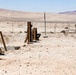 A soldier in desert camouflage uniform is positioned between wooden posts in an arid desert training environment, appearing to be in a tactical kneeling or crouching stance while holding what appears to be a weapon system. The setting shows a military training range with utility poles, sparse desert vegetation, distant structures, and mountain ridgelines in the background. Dust disturbance near the soldier suggests recent movement or a weapon discharge.