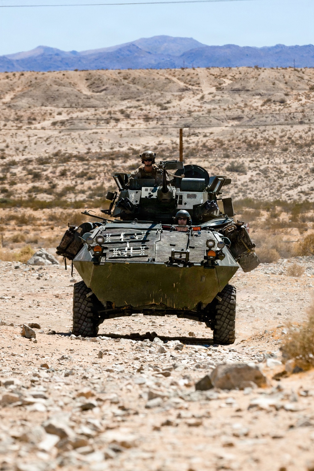 A wheeled armored fighting vehicle, consistent with an LAV-25 (Light Armored Vehicle), is traveling head-on along a rocky desert trail. Two crew members are visible — one in the turret hatch and one in the driver's hatch. The arid, mountainous desert background suggests a training environment such as the Mojave Desert or a similar U.S. military installation.