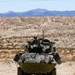 A wheeled armored fighting vehicle, consistent with an LAV-25 (Light Armored Vehicle), is traveling head-on along a rocky desert trail. Two crew members are visible — one in the turret hatch and one in the driver's hatch. The arid, mountainous desert background suggests a training environment such as the Mojave Desert or a similar U.S. military installation.