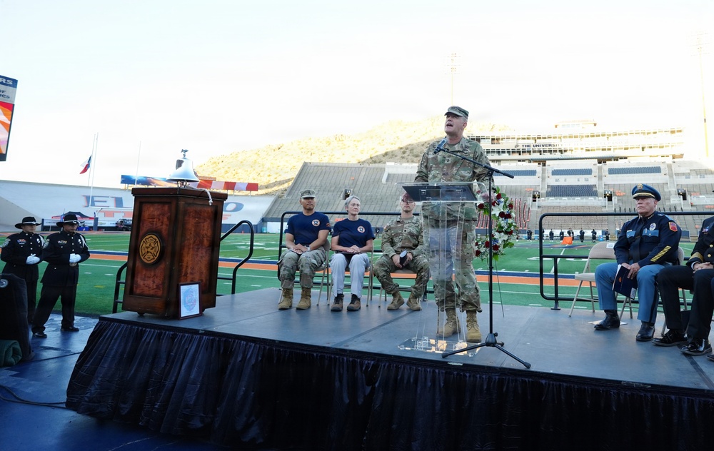 A formal memorial or commemorative ceremony is taking place inside a stadium, likely at the University of Texas at El Paso (UTEP) based on visible signage. A military officer in Army Combat Uniform (ACU) is speaking at a clear podium on a raised stage, flanked by law enforcement, military personnel, and first responders. The presence of a floral wreath, flags at half-staff, and uniformed honor guards suggests this is a solemn remembrance event, possibly a 9/11 memorial.