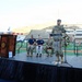 A formal memorial or commemorative ceremony is taking place inside a stadium, likely at the University of Texas at El Paso (UTEP) based on visible signage. A military officer in Army Combat Uniform (ACU) is speaking at a clear podium on a raised stage, flanked by law enforcement, military personnel, and first responders. The presence of a floral wreath, flags at half-staff, and uniformed honor guards suggests this is a solemn remembrance event, possibly a 9/11 memorial.