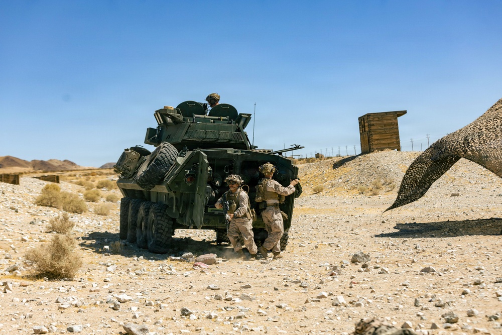 The image shows military personnel in desert camouflage uniforms operating alongside a wheeled armored vehicle, likely an LAV-25 (Light Armored Vehicle), in an arid desert training environment. Two soldiers are visible on the ground near the vehicle's side door while a third is positioned in the turret hatch on top. The setting appears to be a military training range based on the constructed wooden structures and camouflage netting visible in the background.