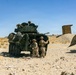 The image shows military personnel in desert camouflage uniforms operating alongside a wheeled armored vehicle, likely an LAV-25 (Light Armored Vehicle), in an arid desert training environment. Two soldiers are visible on the ground near the vehicle's side door while a third is positioned in the turret hatch on top. The setting appears to be a military training range based on the constructed wooden structures and camouflage netting visible in the background.