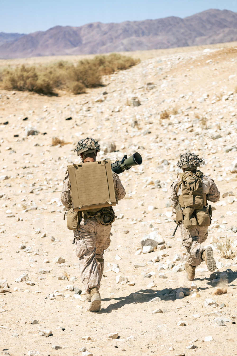Two military personnel in desert camouflage uniforms are moving through an arid, rocky desert terrain. The lead soldier is carrying what appears to be a shoulder-fired rocket launcher (likely an AT4 or similar recoilless weapon) along with a large tan equipment case on their back. The background features sparse desert vegetation, rocky ground, and distant mountain ridges under a clear sky.
