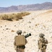 Two military personnel in desert camouflage uniforms are moving through an arid, rocky desert terrain. The lead soldier is carrying what appears to be a shoulder-fired rocket launcher (likely an AT4 or similar recoilless weapon) along with a large tan equipment case on their back. The background features sparse desert vegetation, rocky ground, and distant mountain ridges under a clear sky.