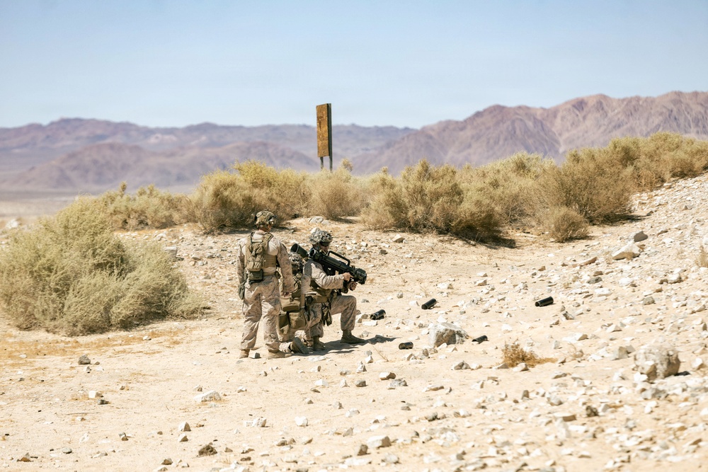 Two military personnel in desert camouflage uniforms are positioned in an arid desert training environment. One soldier is kneeling and aiming a large shoulder-mounted weapon system, while the other stands nearby in a support role. The setting appears to be a military training range based on the visible target post and desert terrain.