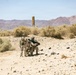Two military personnel in desert camouflage uniforms are positioned in an arid desert training environment. One soldier is kneeling and aiming a large shoulder-mounted weapon system, while the other stands nearby in a support role. The setting appears to be a military training range based on the visible target post and desert terrain.