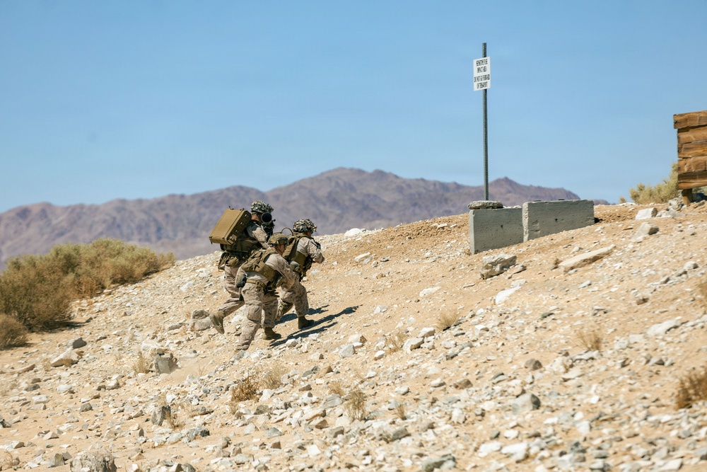 Two military personnel in desert camouflage uniforms are moving tactically up a rocky hillside in an arid desert environment. One soldier is carrying a large equipment pack on their back. The setting appears to be a military training area, evidenced by concrete barriers and a posted sign in the background.