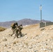 Two military personnel in desert camouflage uniforms are moving tactically up a rocky hillside in an arid desert environment. One soldier is carrying a large equipment pack on their back. The setting appears to be a military training area, evidenced by concrete barriers and a posted sign in the background.