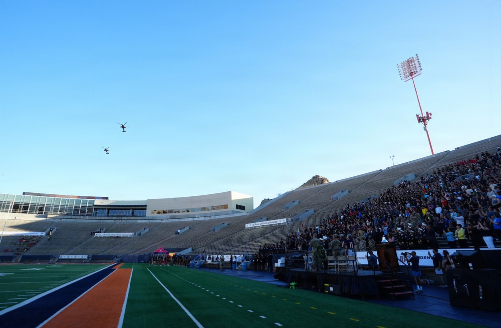 A military or ceremonial event is taking place at an American football stadium, with two helicopters performing a flyover above the field. The stadium is partially filled with spectators, and military personnel are visible on the field near a podium. The scene appears to be a formal ceremony, possibly a commissioning, graduation, or pre-game military tribute.