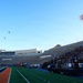 A military or ceremonial event is taking place at an American football stadium, with two helicopters performing a flyover above the field. The stadium is partially filled with spectators, and military personnel are visible on the field near a podium. The scene appears to be a formal ceremony, possibly a commissioning, graduation, or pre-game military tribute.