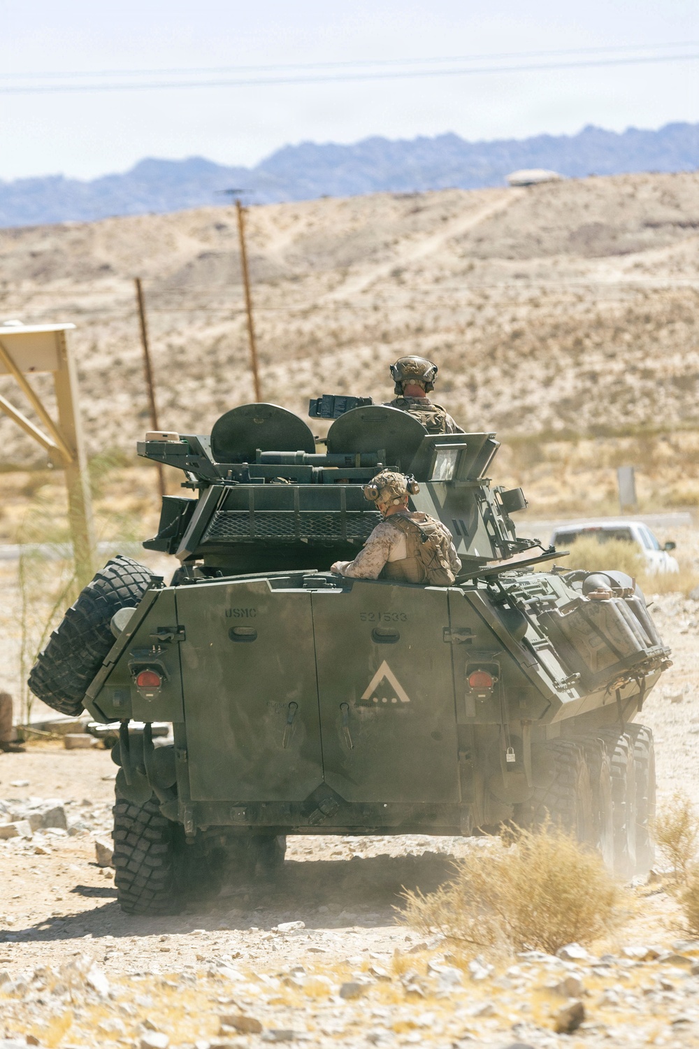 A USMC (United States Marine Corps) Light Armored Vehicle (LAV) is moving through a desert terrain, likely during a training exercise. Two Marines are visible in the open turret hatches, wearing combat helmets and tactical gear. The arid, mountainous landscape in the background is consistent with a Southwestern U.S. military training installation.