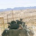 A USMC (United States Marine Corps) Light Armored Vehicle (LAV) is moving through a desert terrain, likely during a training exercise. Two Marines are visible in the open turret hatches, wearing combat helmets and tactical gear. The arid, mountainous landscape in the background is consistent with a Southwestern U.S. military training installation.