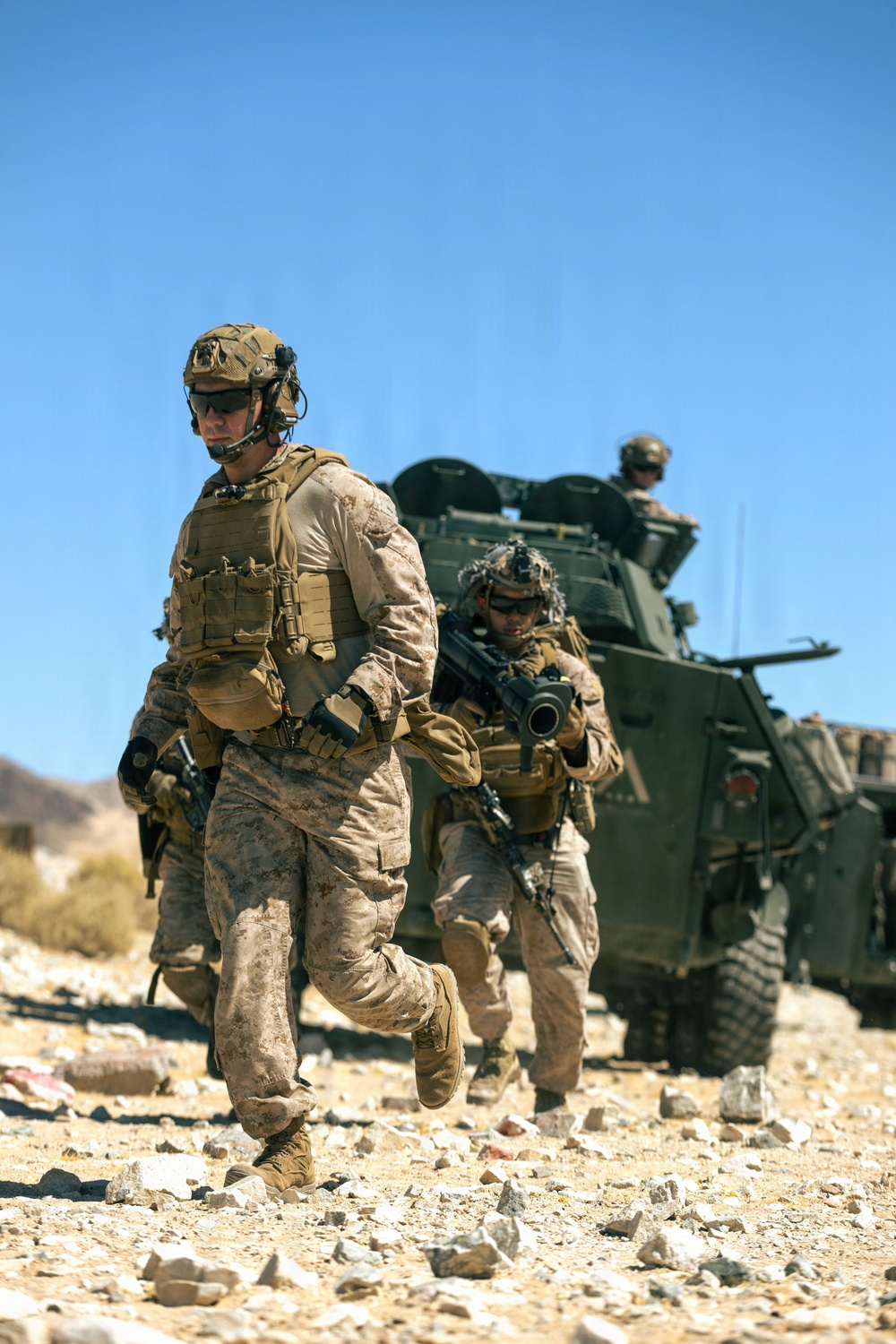 Two dismounted soldiers in desert camouflage (MARPAT pattern) move on foot across rocky, arid terrain while a third occupant mans a position atop an armored vehicle in the background. The scene depicts a tactical dismount operation in a desert environment under clear blue skies. Equipment and uniforms are consistent with U.S. Marine Corps gear and doctrine.
