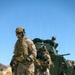 Two dismounted soldiers in desert camouflage (MARPAT pattern) move on foot across rocky, arid terrain while a third occupant mans a position atop an armored vehicle in the background. The scene depicts a tactical dismount operation in a desert environment under clear blue skies. Equipment and uniforms are consistent with U.S. Marine Corps gear and doctrine.