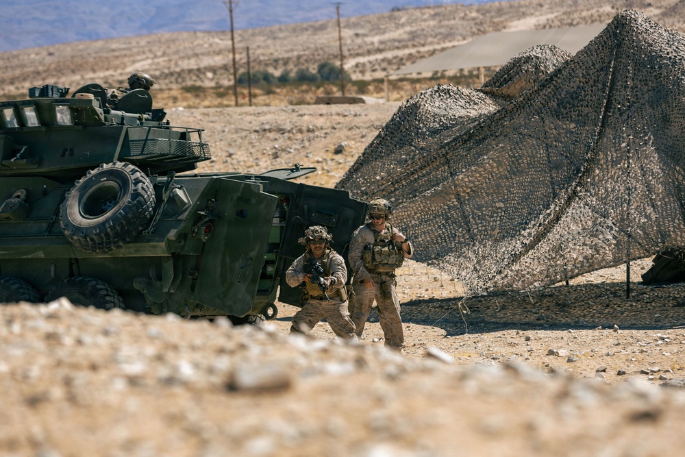 Two armed military personnel in desert camouflage uniforms and tactical gear are positioned beside a large armored wheeled vehicle, likely an LAV (Light Armored Vehicle), in an arid desert environment. A camouflage netting structure is erected nearby, suggesting a forward operating position or field exercise. The setting appears to be a desert training area based on the terrain and infrastructure visible in the background.