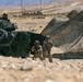 Two armed military personnel in desert camouflage uniforms and tactical gear are positioned beside a large armored wheeled vehicle, likely an LAV (Light Armored Vehicle), in an arid desert environment. A camouflage netting structure is erected nearby, suggesting a forward operating position or field exercise. The setting appears to be a desert training area based on the terrain and infrastructure visible in the background.