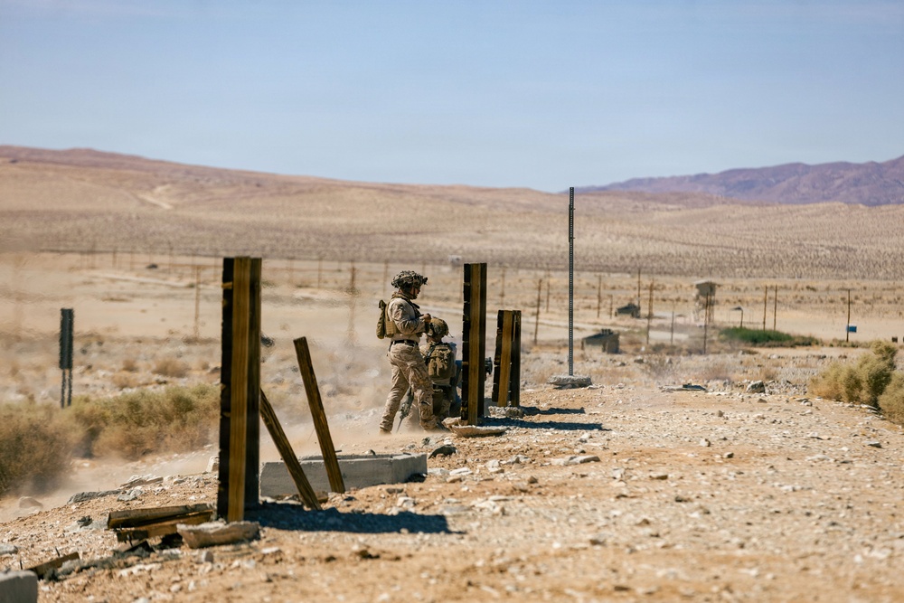 A soldier in desert camouflage uniform and tactical gear is operating what appears to be a mortar system in an arid desert training or combat environment. The scene shows a sparse, rocky desert landscape with scattered vertical metal posts and deteriorating concrete structures. The background reveals an expansive desert valley with distant mountains under a clear sky.