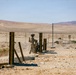 A soldier in desert camouflage uniform and tactical gear is operating what appears to be a mortar system in an arid desert training or combat environment. The scene shows a sparse, rocky desert landscape with scattered vertical metal posts and deteriorating concrete structures. The background reveals an expansive desert valley with distant mountains under a clear sky.