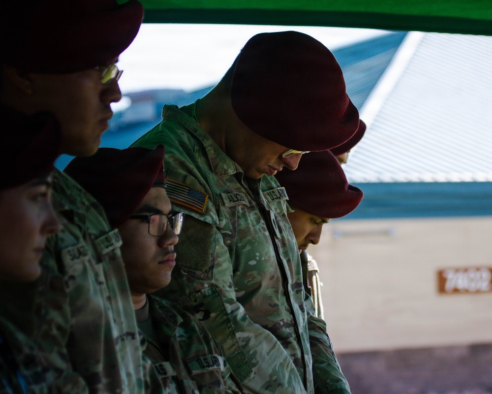 A group of U.S. Army soldiers wearing maroon berets and OCP (Operational Camouflage Pattern) uniforms are standing in a line with heads bowed, suggesting a moment of prayer or reflection. The soldiers appear to be at an outdoor military ceremony or formation, partially sheltered under a green canopy. American flag patches are visible on their uniforms, identifying them as U.S. Army personnel.