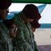 A group of U.S. Army soldiers wearing maroon berets and OCP (Operational Camouflage Pattern) uniforms are standing in a line with heads bowed, suggesting a moment of prayer or reflection. The soldiers appear to be at an outdoor military ceremony or formation, partially sheltered under a green canopy. American flag patches are visible on their uniforms, identifying them as U.S. Army personnel.